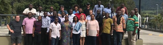 A group of around 30 people standing in the sun during the CORDEX Africa workshop 2018, in the background there are trees and green mountains surrounded by clouds.